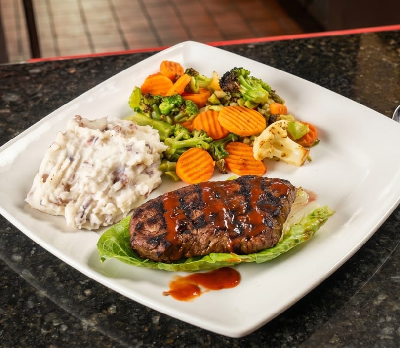 Grilled steak with BBQ sauce, mashed potatoes, and mixed vegetables on a white plate.